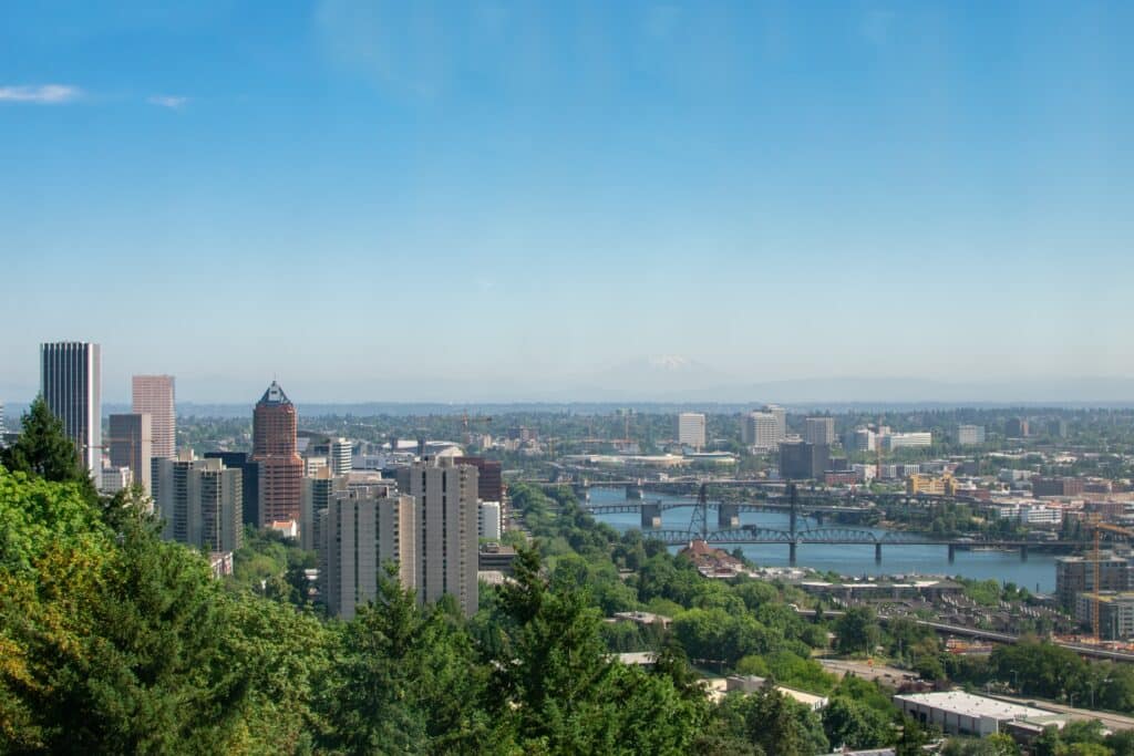 A landscape shot of Portland's skyline. The Willamette River is visible in frame, as are a smattering of skyscrapers on the left hand side of the screen. Trees pepper each area.